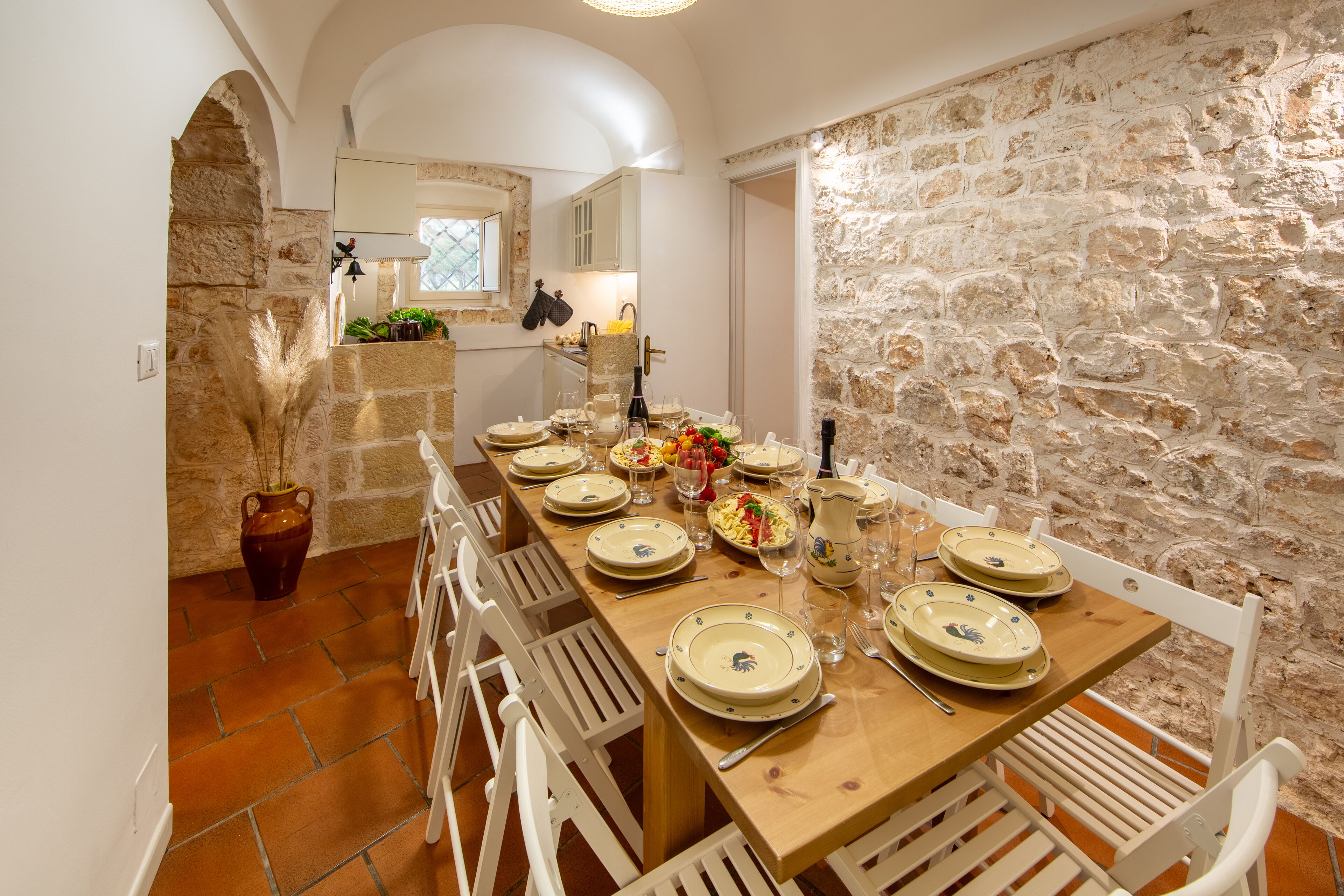 Un'area pranzo con parete in pietra, tavolo in legno apparecchiato per sei persone, sedie bianche e una pianta in vaso.