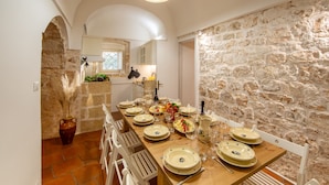 Un coin repas avec un mur en pierre, une table en bois dressée pour six personnes, des chaises blanches et une plante en pot.