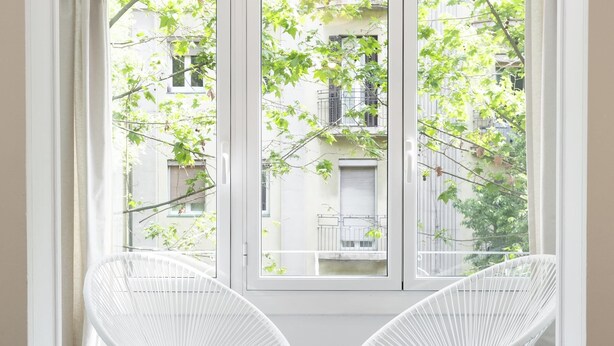 Ein Zimmer mit zwei weißen Korbstühlen, die zu einem Fenster mit Blick auf ein Gebäude mit Balkonen blicken.