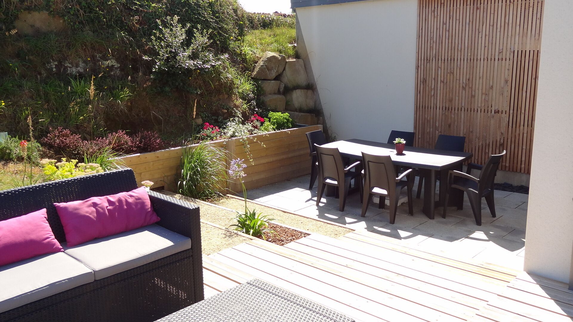 Una terrazza esterna con tavolo e sedie in legno, un divano in vimini con cuscini, e diverse piante e fiori.