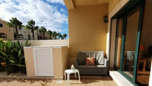 Un balcon avec un canapé et une petite table, un mur blanc et une vue sur des palmiers et des immeubles.