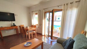 Un salon comprenant un canapé, une table basse, un téléviseur, un coin repas avec des chaises et une table, ainsi qu’une porte-fenêtre coulissante donnant sur une piscine extérieure.