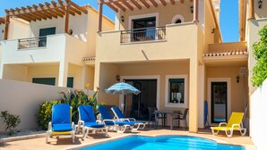 Un espace piscine avec des chaises longues et des parasols, une terrasse couverte et un bâtiment doté de balcons.