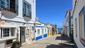 Une rue étroite, bordée d’immeubles peints en blanc et en bleu, un café avec des tables en terrasse, et une vue dégagée sur la mer au loin.