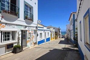 A narrow street with buildings painted in white and blue, a café with outdoor seating, and a clear view of the sea in the distance. - DomusIberica Burgau, 2 bedroom house with A/C, private pool, walk to the beach! (Burgau)