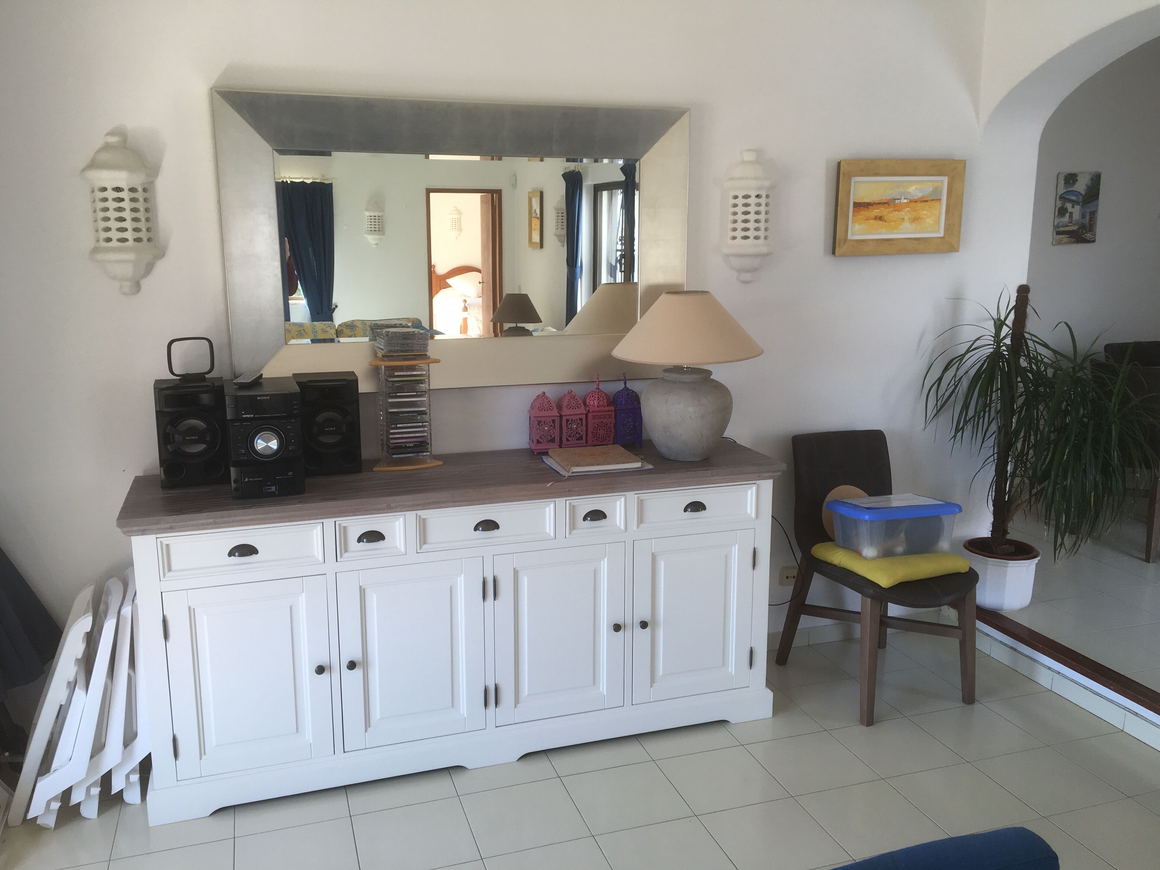 A room with a white cabinet, a mirror, a lamp, a plant, and a chair.