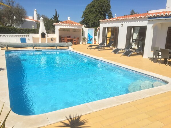 A rectangular swimming pool with a tiled edge, surrounded by lounge chairs and a covered seating area.