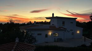 Ein weißes Gebäude mit einem Ziegeldach und einem kleinen Turm, umgeben von Bäumen und einem Abendhimmel.
