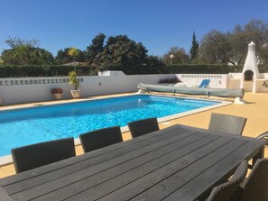 A swimming pool with a tiled edge, surrounded by a white wall with decorative trim. There are lounge chairs and a table with chairs in the foreground.