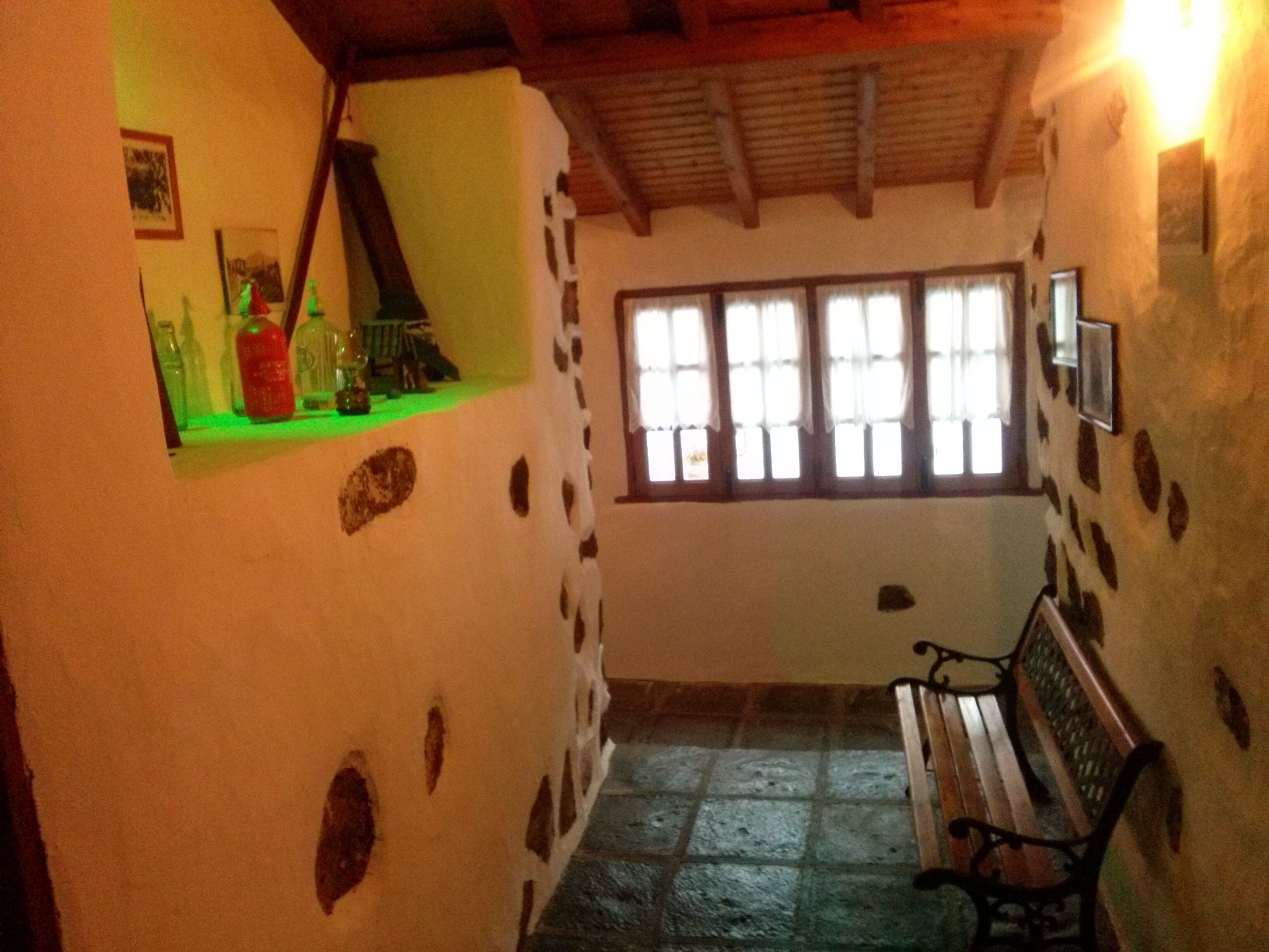 A room with a bench, a window with curtains, and a shelf with bottles.