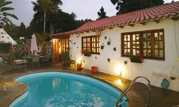 Zona junto a la piscina con piscina, patio con mobiliario exterior y edificio con tejado de tejas y ventanas.