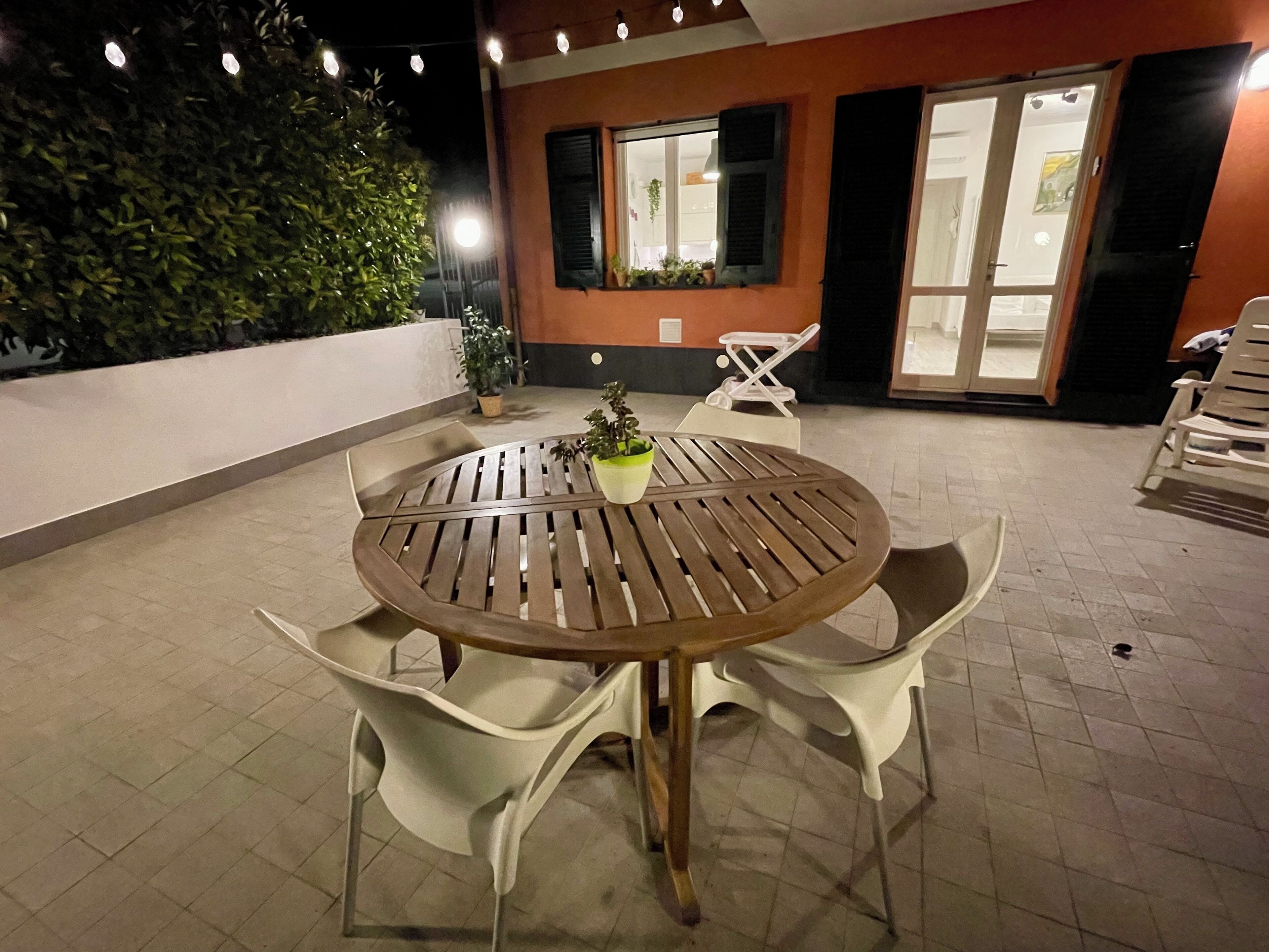 Eine Terrasse im Freien mit einem runden Holztisch und vier weißen Plastikstühlen. Auf dem Tisch steht ein Blumentopf. Die Terrasse grenzt an ein Gebäude mit Glastür und Fenstern.