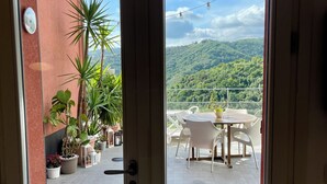 Blick durch eine Glastür auf eine Terrasse mit Tisch und Stühlen, Blumentöpfen und einer Berglandschaft im Hintergrund.