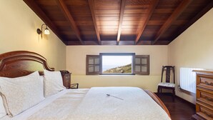 A bedroom with a wooden ceiling, a large bed with patterned bedding, a nightstand, a dresser, and a window with a view of a landscape. - El Nogal House, La Orotava (La Orotava)