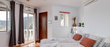A bedroom with a bed, curtains, an air conditioning unit, a ceiling fan, and a view through the door.