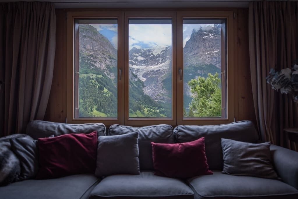 A room with a sofa facing a large window with a mountain view.