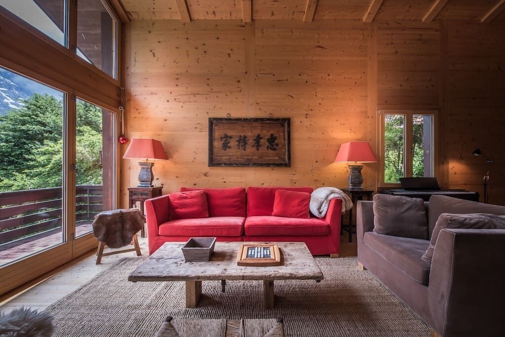 Un salon chaleureux avec un canapé rouge, une table basse en bois et deux lampes. Une œuvre d’art encadrée est accrochée au mur, et on peut admirer la verdure à travers la fenêtre.