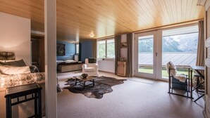 A spacious room with a wooden ceiling, a large window offering a mountain view, a cozy seating area with a white chair and a small table, and a carpeted floor. - Cozy free standing Chalet with garden and amazing mountain views (Grindelwald)