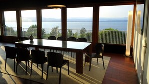 Un comedor con una mesa grande y sillas, ventanas amplias con vista al mar y a las montañas, y un balcón con barandilla.