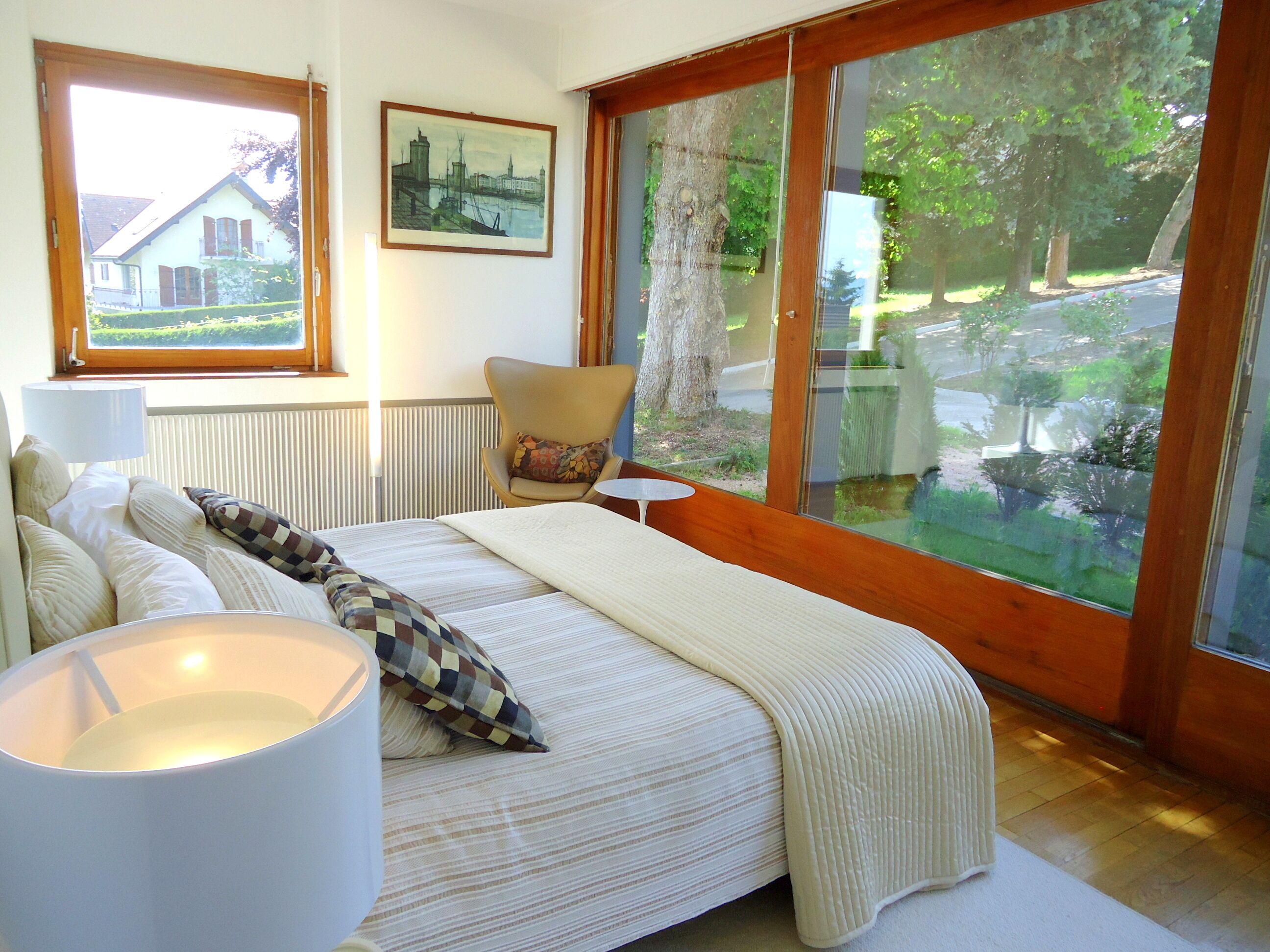Ein Schlafzimmer mit einem großen Bett, einem Sessel, einem Tisch und einem Fenster mit Blick auf ein Haus und Bäume.
