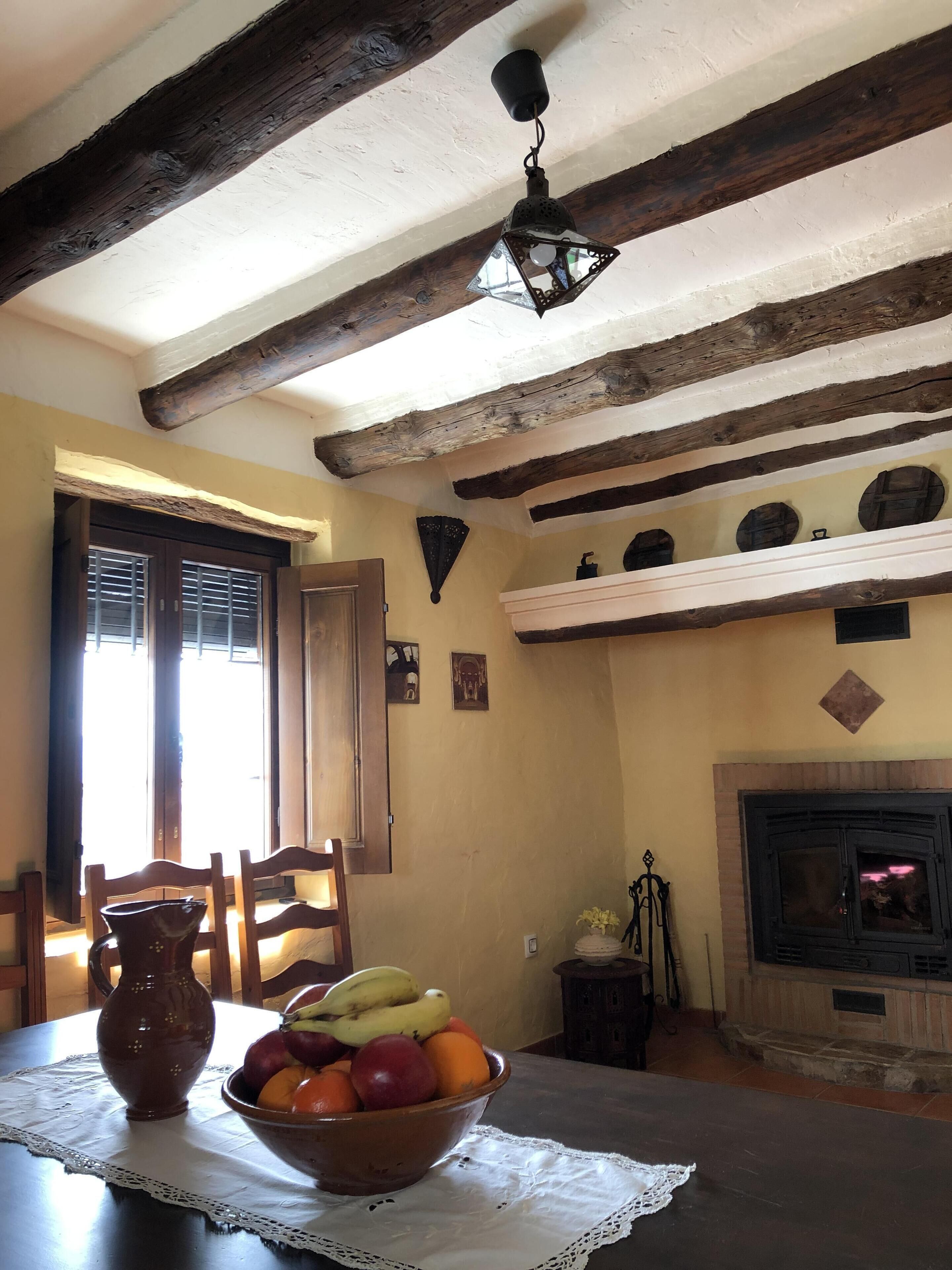 A rustic interior with exposed wooden beams, a fireplace, and a table with a fruit bowl.