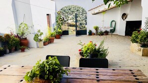 An outdoor patio area with a wooden table and chairs, surrounded by potted plants and a view of a garden through an archway.