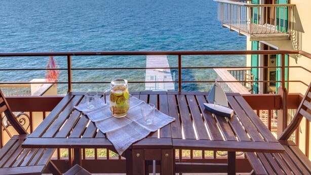 Une table en bois, sur laquelle se trouvent une carafe de limonade et des verres, donnant sur une mer avec des montagnes en arrière-plan.