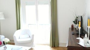 Ett ljust rum med en vit stol, ett litet bord med en vas och ett skrivbord i trä med en växt och ljus.