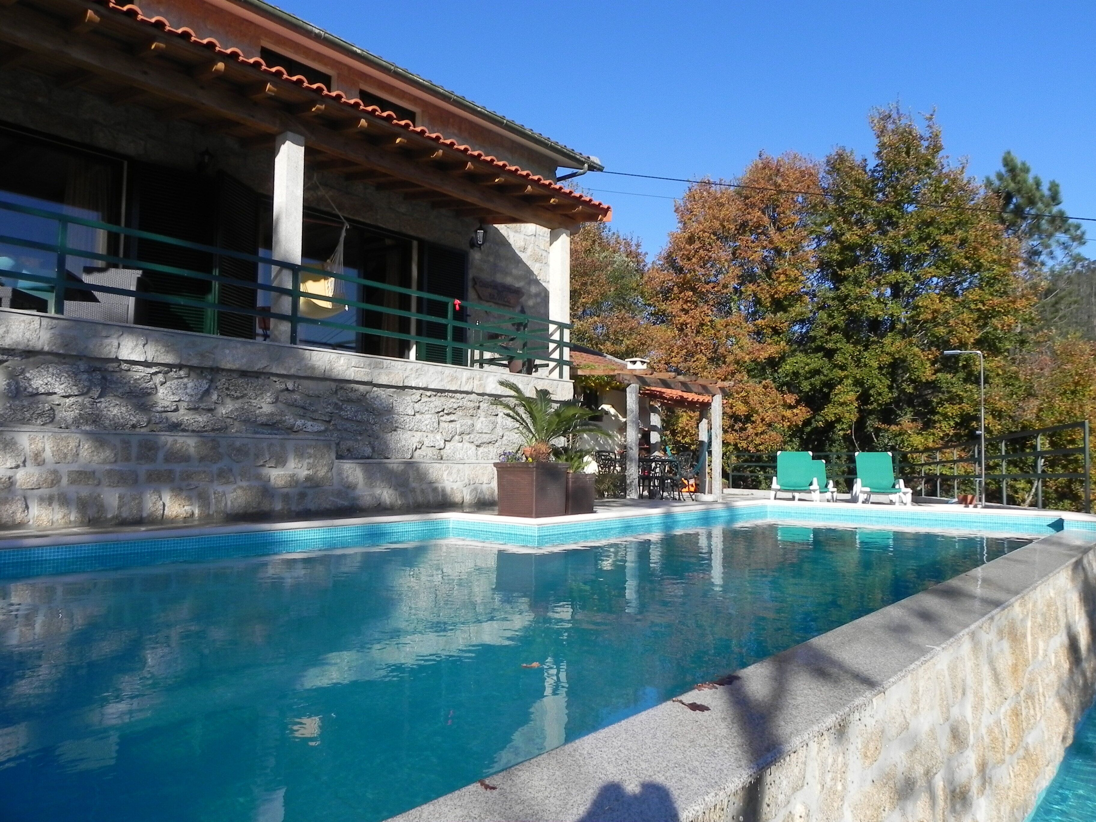A swimming pool with a stone wall, a covered seating area, and a building with a tiled roof.