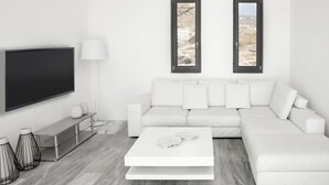 Una sala de estar moderna con un sofá blanco, un televisor de pantalla plana, una mesa de centro y dos ventanas con vistas a un paisaje rocoso.
