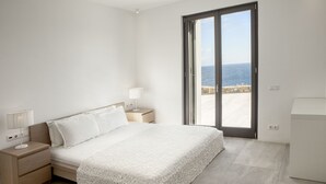 Un dormitorio con cama, mesitas de noche, lámpara y vista al mar a través de un ventanal corredero.