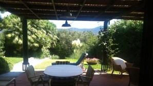 Un patio techado al aire libre con una mesa redonda y sillas, rodeado de vegetación exuberante y vistas a montañas distantes.