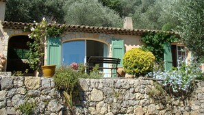 Un mur de pierres rustique, avec une maison en pierre dotée de volets verts, d’une grande fenêtre et d’un petit balcon.