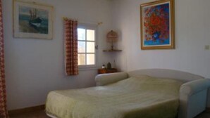 Une chambre à coucher avec un lit, une fenêtre avec des rideaux, un tableau et une petite étagère sur laquelle est posé un objet décoratif.