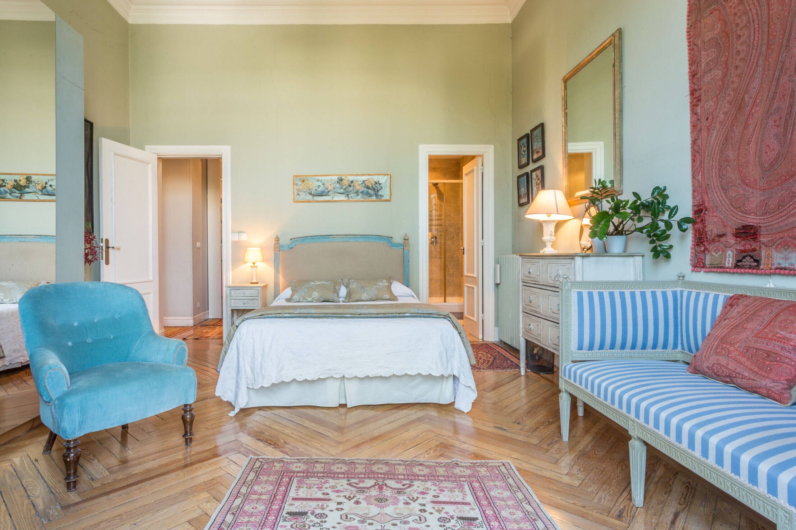 Une chambre à coucher avec un lit, un fauteuil bleu, un canapé à rayures, une table de chevet avec une lampe, une commode et un miroir.