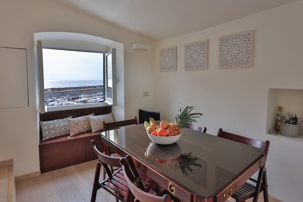 A dining area with a view of the sea, a wooden bench with cushions, a table with a fruit bowl, and decorative wall art.