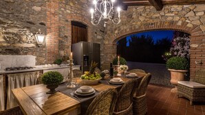 Un área de comedor al aire libre con paredes de piedra, una mesa de madera dispuesta para cuatro personas, sillas de mimbre y una araña.