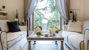Habitación con sofá blanco, una mesa de centro con objetos decorativos y vistas a follaje a través de cortinas translúcidas.