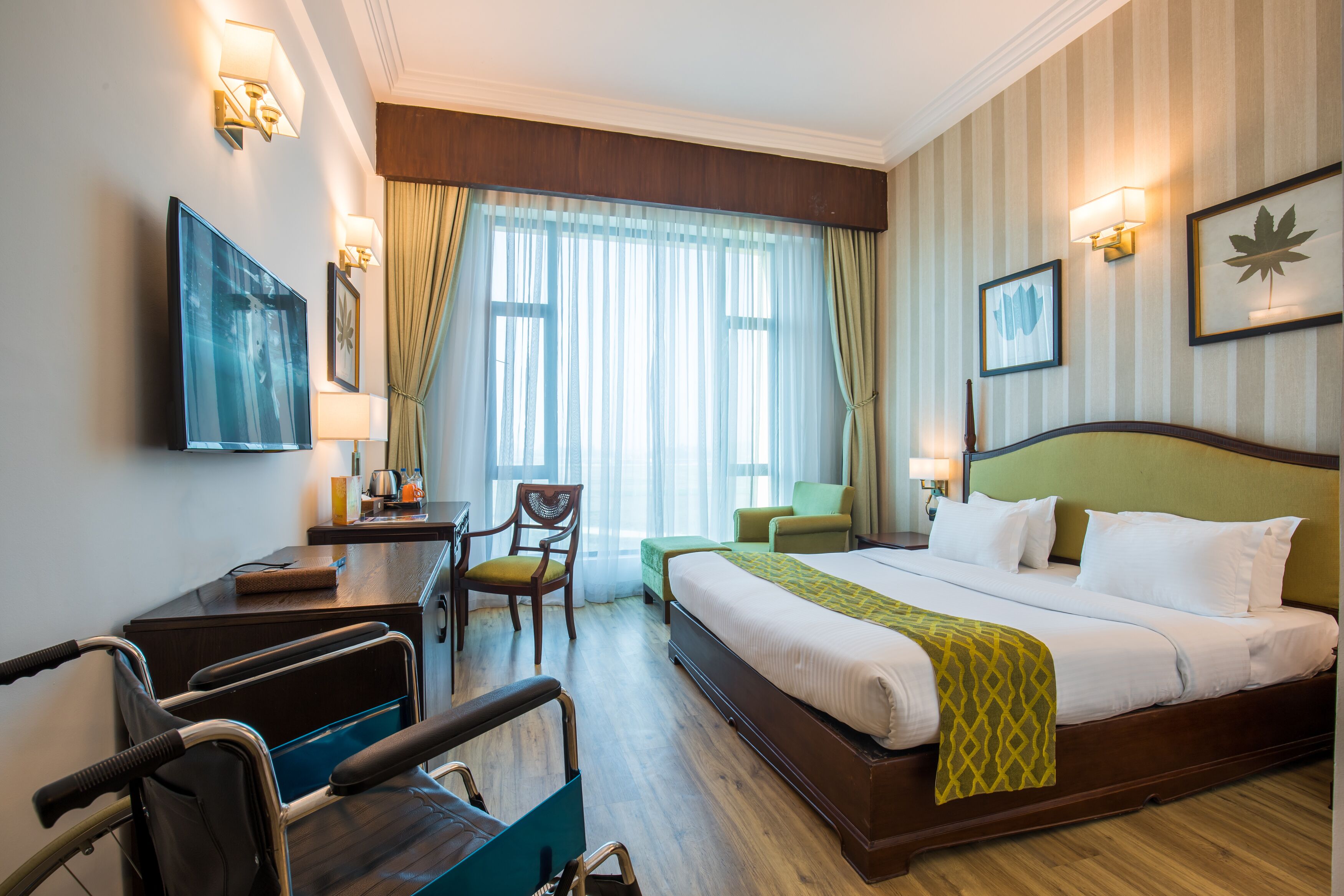 A hotel room with a large bed, a desk with a chair, a wheelchair, and a TV on the wall.