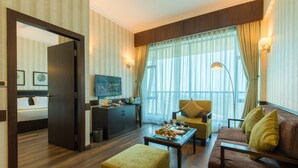A hotel room with a wooden floor, a sofa, a coffee table with fruit, a TV, and a bed in the adjacent room. - Soaltee Westend Premier (Nepalgunj)