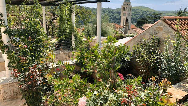 Un jardín con pérgola, muros de piedra y vista a una torre de iglesia.