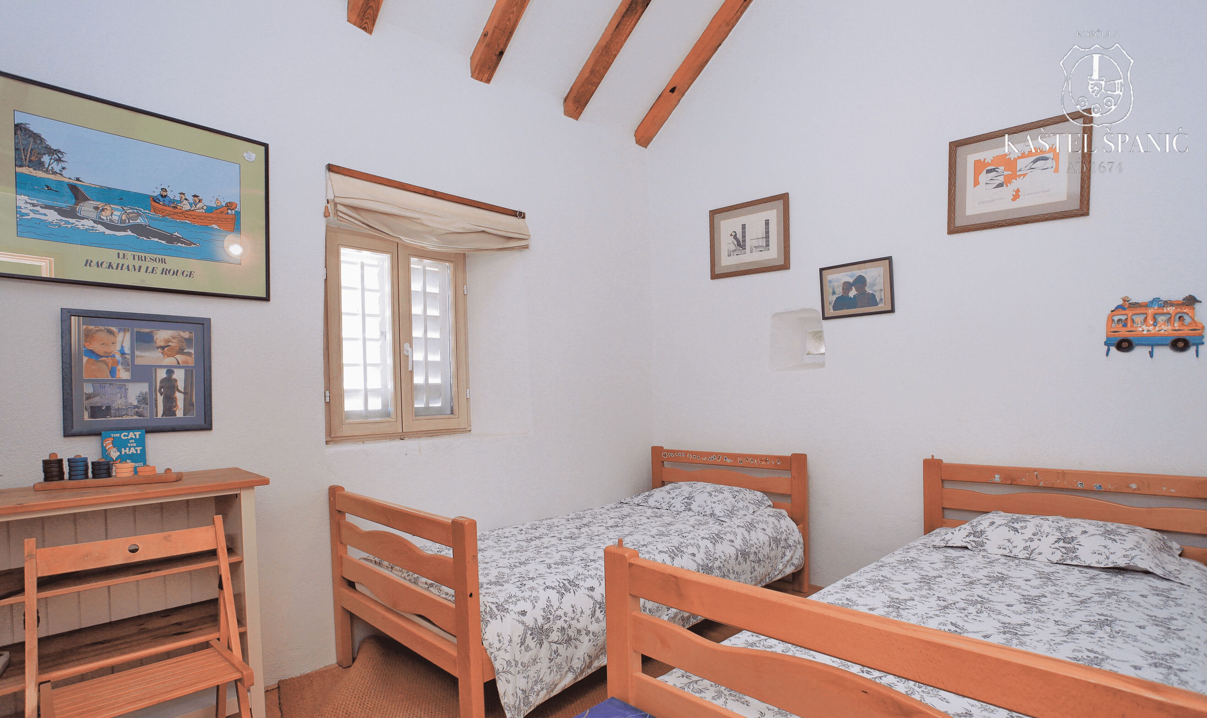 A room with two wooden beds, a desk, and a window with curtains. There are framed pictures on the walls.