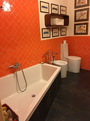 A bathroom with a white bathtub, a white sink, and a toilet. There are framed pictures on the wall and a shelf with a towel. - Villa with heated pool (Castelbuono (Pa))
