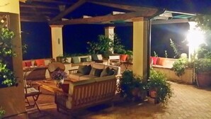Un área de estar al aire libre con un banco y sillas de madera, rodeada de plantas en macetas y una estructura de pérgola.