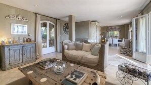 A spacious living area with a large sofa, a coffee table with books and decorative items, a dining area with a table and chairs, and a kitchen visible through an open door. - Villa La Lavande Has A Heated Pool And Magnificent Sea, Golf And Mountain Views (Châteauneuf-Grasse)