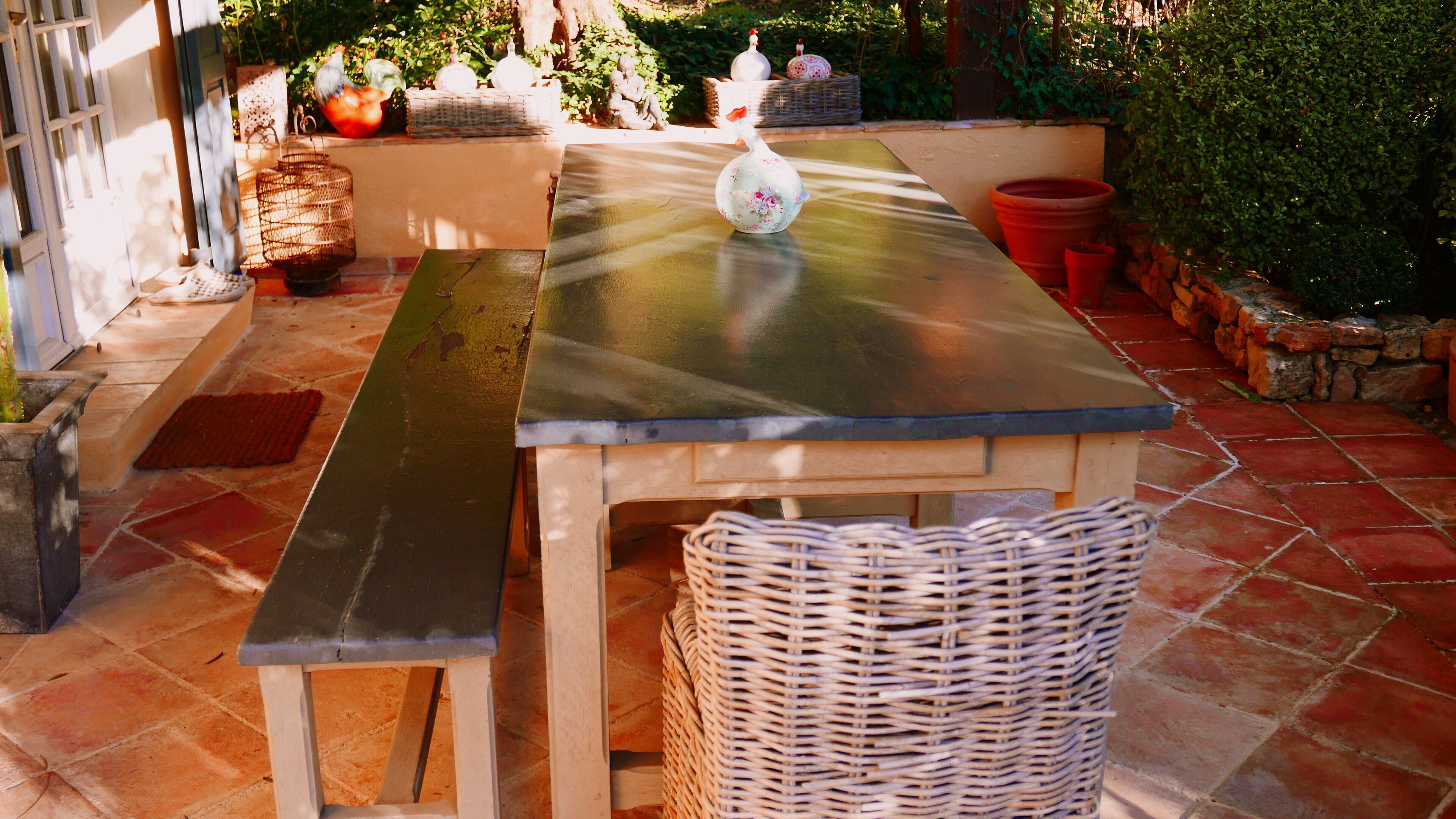 Um pátio com mesa e bancos de metal, rodeado por plantas em vasos e uma cesta de vime.