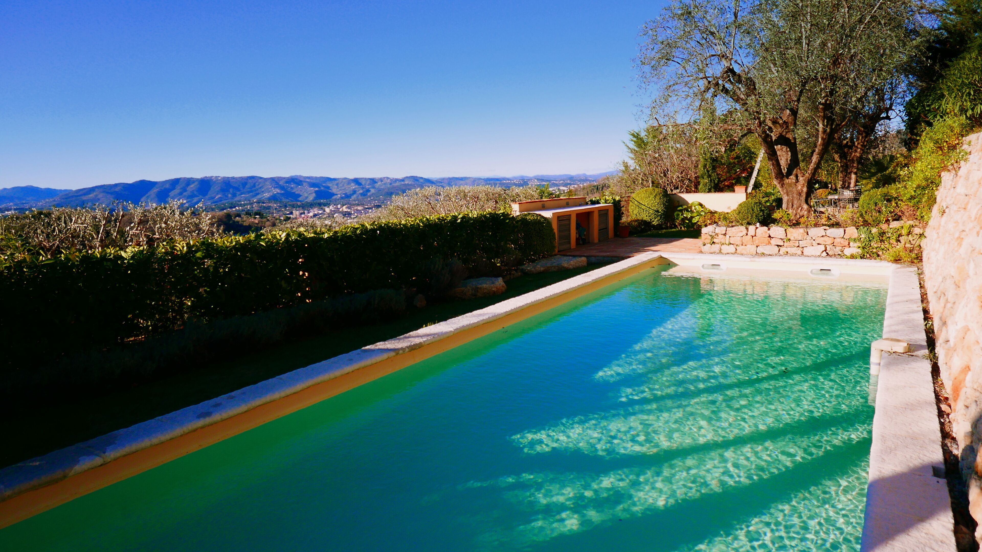 Ein Swimmingpool mit klarem Wasser, umgeben von Steinmauern und Grünpflanzen, mit Blick auf entfernte Berge.