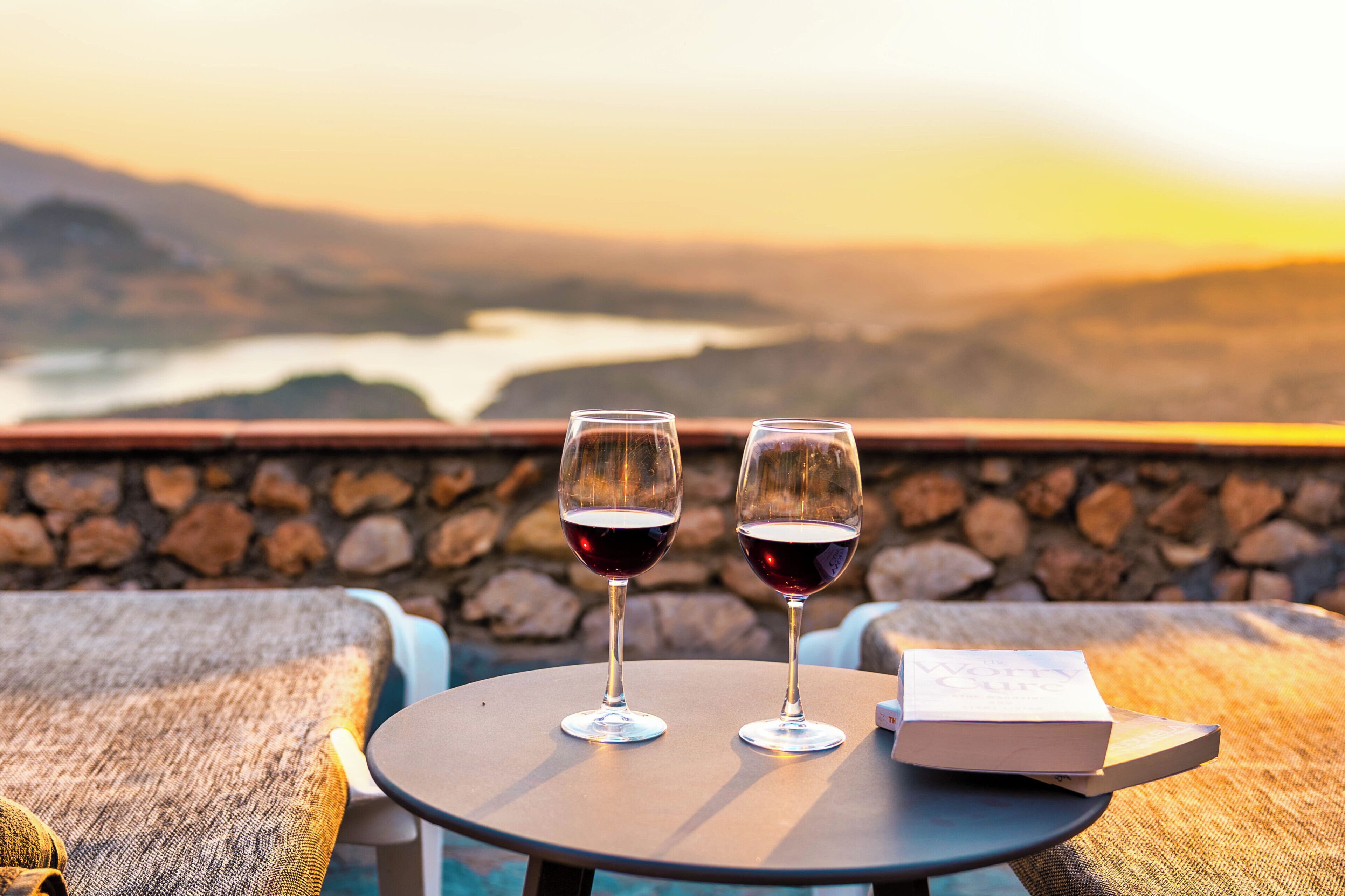 Two wine glasses on a table with a book beside them, overlooking a scenic view of mountains and a body of water.