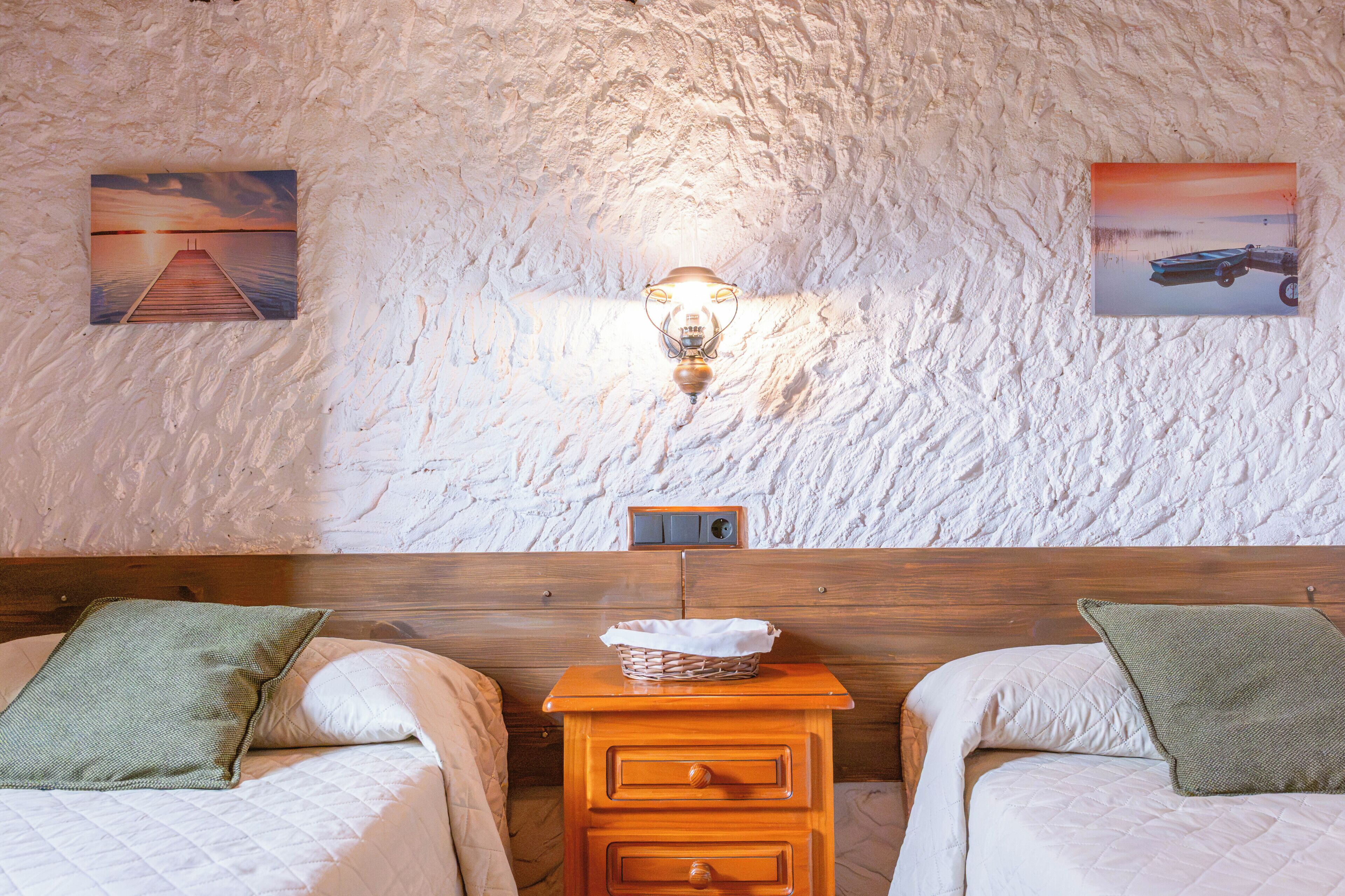 Una habitación con dos camas, una mesita de noche de madera, una lámpara de pared y dos cuadros enmarcados.