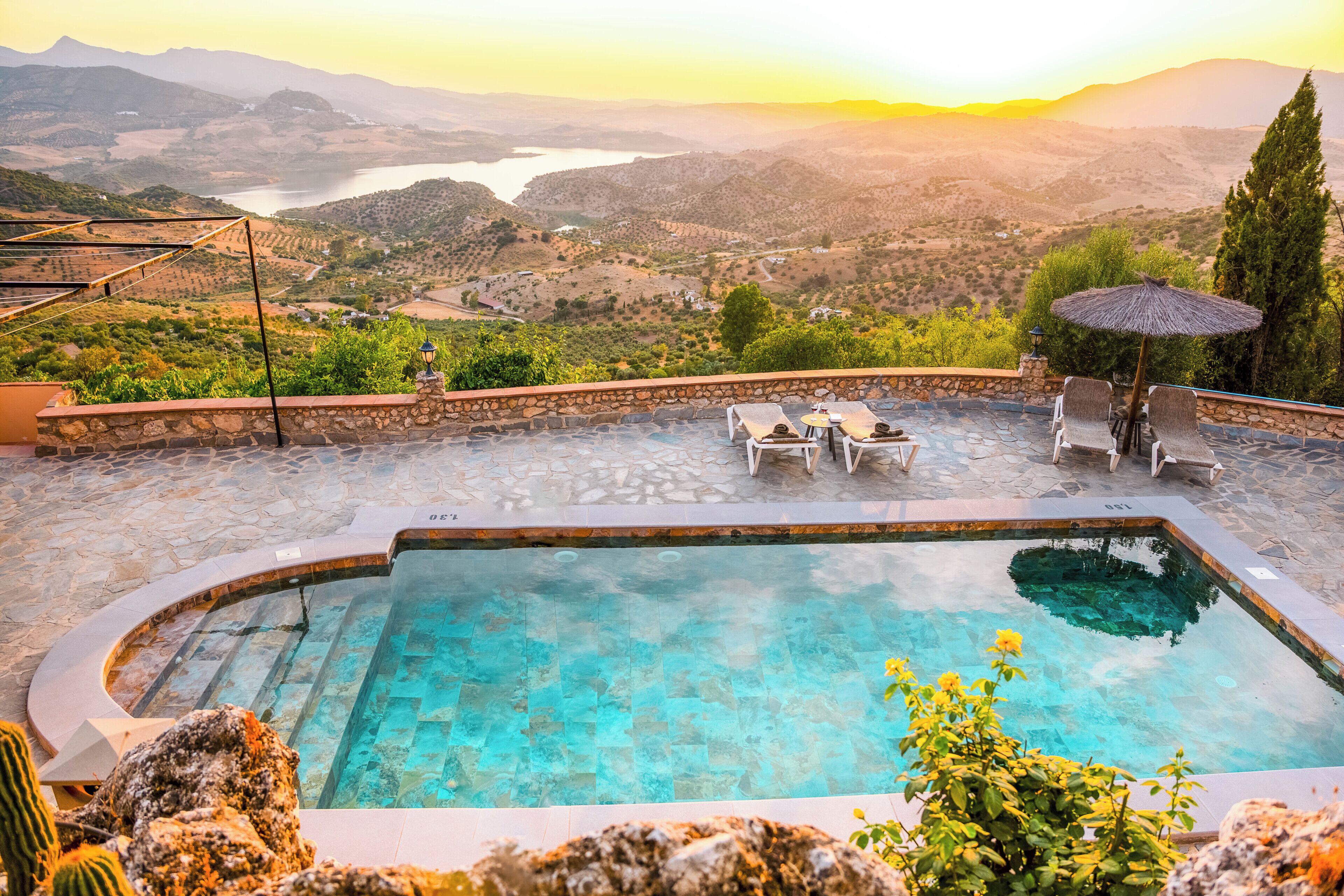 A swimming pool with a stone border, surrounded by lounge chairs and a table, overlooking a mountainous landscape with a view of a lake and hills.